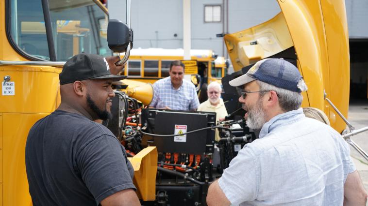 Two people stand and speak near an open electric school bus hood.