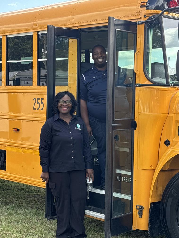 Two people stand at the entrance of an electric school bus, smiling.  