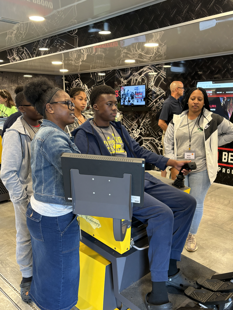 A young person sits in a machinery simulator chair, surrounded by other young people. 