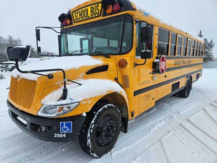 A wheelchair accessible Shawnee Public Schools electric school bus is parked, covered with some snow, standing on a snowy road.