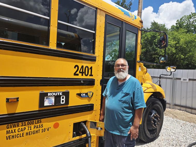 Ron Reser stands next to the electric school bus he drives for Shawnee Public Schools. While each of Shawnee’s school buses has an animal printout for identification, his is the only one in the district with a personalized character — a gift from his co-workers in recognition of his soft demeanor. Image by Thomas Wondrock