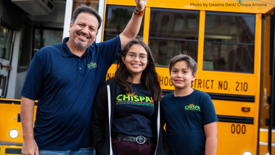 Three people stand in front of an electric school bus smiling as one raises his fist.