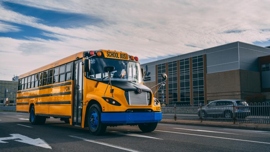 An electric school bus drives near a school.