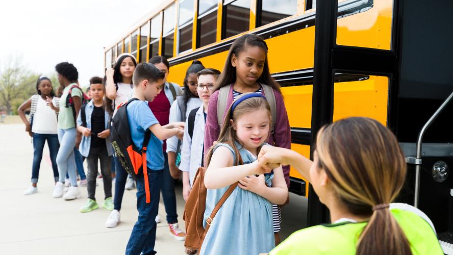 An elementary age girl with Down Syndrome gives a fist bump to a female school bus driver. The girl and other students are boarding the school bus.