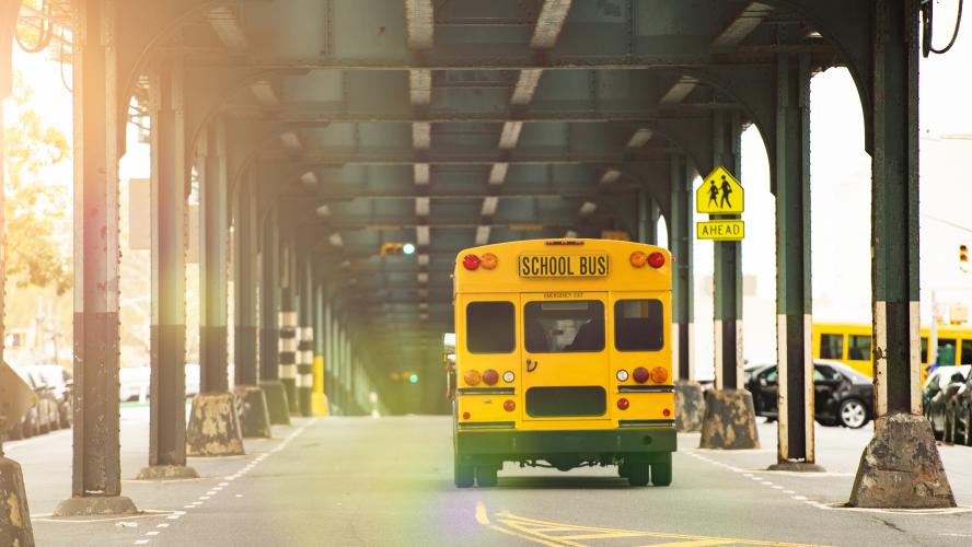 A yellow school bus drives down a road underneath a bridge.