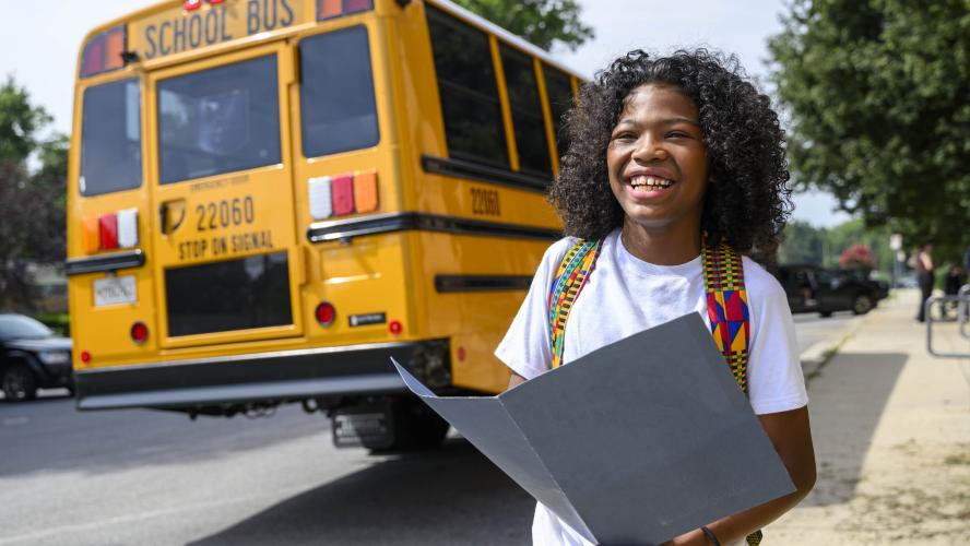 A student holds a folder near an electric school bus and smiles.